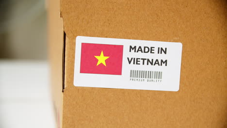 Manos-Aplicando-La-Etiqueta-De-La-Bandera-Hecha-En-Vietnam-En-Una-Caja-De-Cartón-De-Envío-Con-Productos