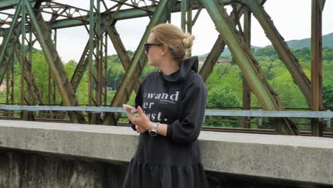 Mädchen-Mit-Brille-Und-Schwarzem-Pullover,-Das-Telefon-überprüft,-Mit-Eiserner-Eisenbahnbrücke-Im-Hintergrund