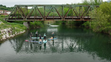 Fly-in-drone-shot-of-people-paddle-boarding-with-big-and-old-iron-train-bride-in-background-and-green-nature-on-sides