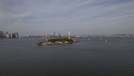 Aerial-view-towards-the-Liberty-island,-surrounded-by-ferries-and-sailboats,-in-sunny-New-York,-USA---rising,-drone-shot