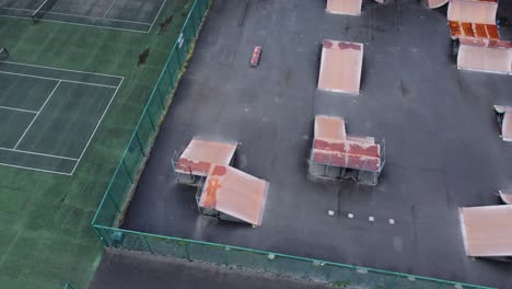 Aerial-view-flying-across-fenced-tennis-court-and-skate-park-ramp-in-empty-closed-playground