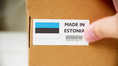 Hands-applying-MADE-IN-ESTONIA-flag-label-on-a-shipping-cardboard-box-with-products