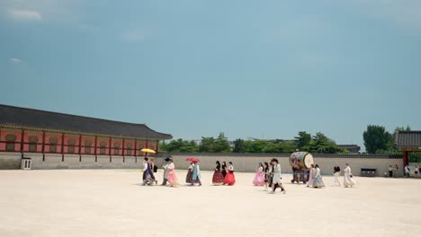 Gruppenreise:-Menschen-In-Farbenfrohen-Traditionellen-Koreanischen-Hanbok-Kleidern-Und-Regenschirmen,-Die-An-Einem-Sommertag-Den-Gyeongbokgung-Palastplatz-Entlang-Spazieren