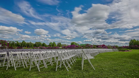 Static-shot-of-rows-of-white-wooden-folding-chairs-on-the-green-grass-surrounded-by-floral-garden-with-white-clouds-passing-by-in-timeplase