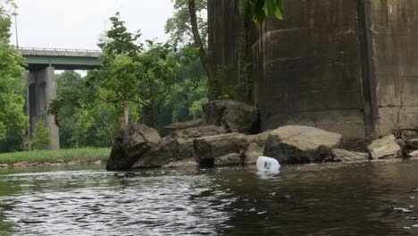 milk-jug-floats-down-river-slowly-towards-old-concrete-bridge-on-a-cloudy-day-wide-panning