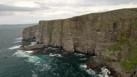 Seabirds-fly-above-a-turquoise-green-ocean-infront-of-a-steep-and-dramatic-cliff-wall-rising-straight-up-out-of-the-ocean-as-waves-crash-against-its-base