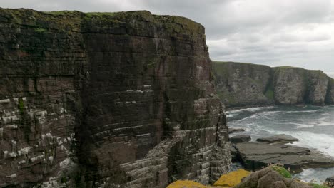 The-camera-slowly-tilts-downwards-to-reveal-a-breathtaking-view-of-a-seabird-colony-with-dramatic-sea-cliffs-as-turquoise-green-waves-crash-against-the-base-of-cliff