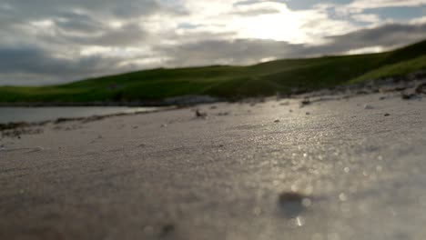 A-low-angle-shot-of-grains-of-golden-sand-on-a-beach-as-the-sun-sets-behind-grassy-hills-and-crashing-waves-in-the-background