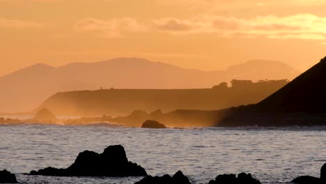 Stunning-golden-sunset-over-sea-at-dusk-with-waves,-ocean-spray-and-layers-of-mountains-in-Wellington,-New-Zealand-Aotearoa