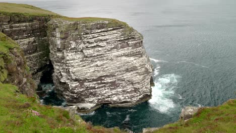 Panning-view-of-a-sea-cliff-and-the-great-sea-stack-of-Handa-Island-covered-in-a-bustling-seabird-colony-full-of-breeding-populations-of-puffins-guillemots,-kittiwake-and-razorbills-in-Scotland