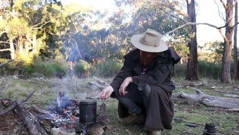 A-bushman-in-an-oil-skin-stockman-jacket-sits-around-a-fire-with-his-billy-boiling-in-the-high-country
