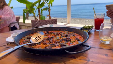 Traditional-Spanish-Paella-dish-with-chicken-and-vegetables-in-a-restaurant-by-the-beach-with-a-sea-view,-eating-green-olives,-tasty-seafood-in-Marbella-Spain,-4K-shot