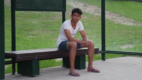 Slow-motion-of-young-Srilankan-man-in-basic-clothes-and-slippers-casually-sitting-and-waiting-at-bus-stop-near-fields