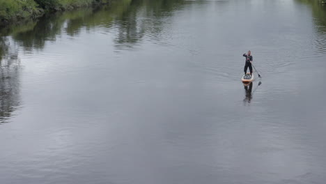 Caucasian-man-in-sunglasses-paddles-paddleboard-on-calm-rural-river