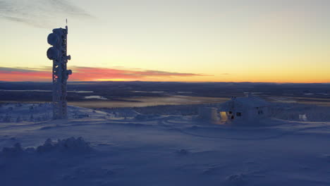 Aerial-view-reversing-across-snow-covered-Lapland-remote-cabin-and-communications-tower-overlooking-polar-wilderness-at-sunrise