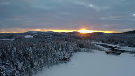 Aerial-view-across-glowing-sunrise-snow-covered-mountains-over-woodland-trees-and-ice-lake-bridge