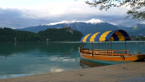 Slow-Panning-Shot-Of-Charming-Cruise-Boat-With-Bled-Lake,-Castle-And-The-Alps-In-The-Background,-Calm-Teal-Pond