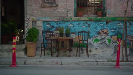 Zwei-Stühle-Und-Ein-Tisch-Vor-Einem-Griechischen-Café-In-Athen-Mit-Straßenkunst-An-Der-Wand