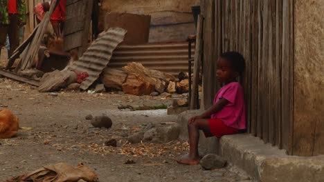 an-african-child-sits-on-the-sill-of-a-dilapidated-house-while-in-the-background-the-animals-walk-around-on-dirty-ground---close-up