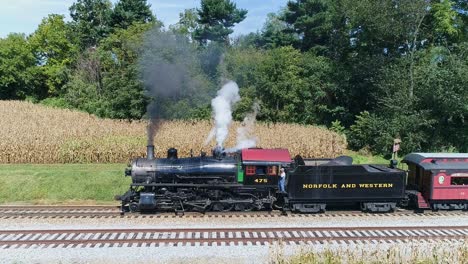 A-Drone-Side-View-of-an-Antique-Steam-Passenger-Train-in-Slow-Motion-Blowing-Smoke-and-Steam-Traveling-While-Stopped-to-Let-Passengers-Off-on-a-Sunny-Summer-Day