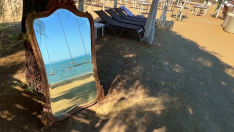 Ungewöhnliche-Aufnahmen-Eines-Spiegels,-Der-An-Einen-Baum-Am-Strand-Gelehnt-Ist-Und-Schaukeln-Und-Im-Meer-Badende-Menschen-Reflektiert