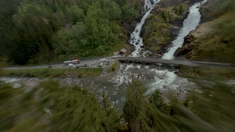 Vuelo-Aéreo-FPV-Cuesta-Abajo-De-Montañas-Verdes-Sobre-Carretera-Con-Tráfico-Y-Cascada-En-Noruega,-LÃ¥tefoss