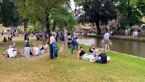 Crowd-of-people-relaxing-in-the-summer-grass-by-the-riverside-in-Bourton-On-The-Water,-Cotswolds---England,-UK