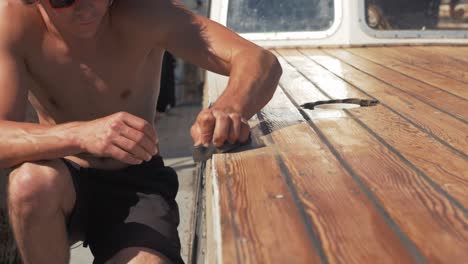 Sanding-roof-planks-of-timber-boat-young-man-blow-dust-off-slow-motion