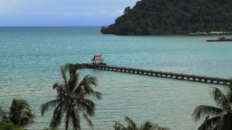 A-pier-with-a-small-red-roof-at-the-end,-in-the-ocean-with-palm-trees-Slowmo