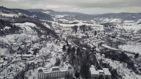 Wide-aerial-shot-of-a-Mining-town-Banska-Stiavnica-in-winter-covered-by-snow,-mountains-in-background,-Cloudy