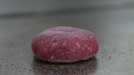 Close-up-of-a-raw-burger-patty-on-a-restaurant-grill-as-black-pepper-is-ground-on-top