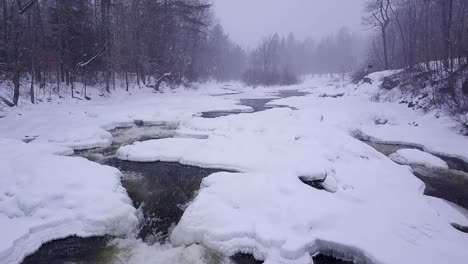 SLOW-MOTION-aerial-shot-flying-upstream-along-an-icy-river-during-a-winter-snow-storm