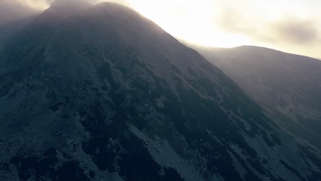 Wide-aerial-drone-slowly-dropping-facing-a-mountain-range-with-heavy-clouds-coming-through-the-mountains