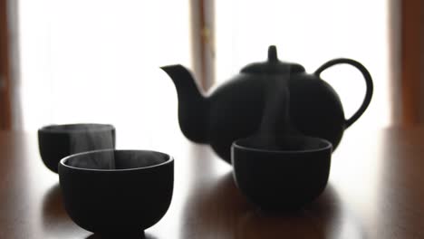 Minimaler-Hintergrund-Eines-Grünen-Japanischen-Teesets-Mit-Dampf,-Der-Aus-Den-Tassen-Kommt,-Auf-Einem-Holztisch,-Mit-Einem-Fenster-Auf-Der-Rückseite
