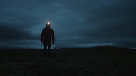Island-Landschaft-Bei-Nacht,-Silhouette-Einer-Person,-Die-Still-Im-Bild-Steht-Und-Mit-Eingeschalteter-Stirnlampe-In-Die-Kamera-Blickt,-Sich-Schnell-Bewegende-Wolken-Im-Hintergrund