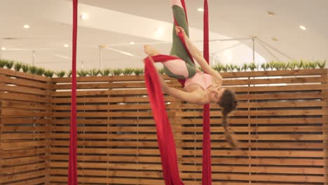 Young-brunette-woman-in-a-minimal-and-modern-gym-with-wooden-dividers-doing-flying-yoga-with-some-bright-red-hammocks,-doing-stunts-and-spinning-around