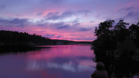 Aerial-drone-view-bypassing-a-man-sitting-at-a-rocky-shore,-of-a-lake,-a-purple-sky,-at-a-colorful-sunset-or-dusk,-at-Albysjon,-Tyreso,-Sweden
