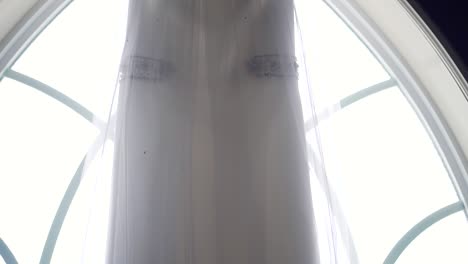 Slow-pan-of-the-top-of-a-wedding-dress-as-it-hangs-from-a-large-window-sill-in-an-old-church
