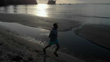 Aerial-tracking-shot-of-silhouetted-young-man-jogging-on-a-beach-in-Auckland,-New-Zealand