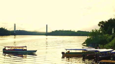 Traditionelles-Langboot-Driftet-Zusammen-Mit-Anderen-Angedockten-Langbooten-An-Die-Küste,-Im-Hintergrund-Die-Brücke-Von-Brasilien-Nach-Französisch-Guayana
