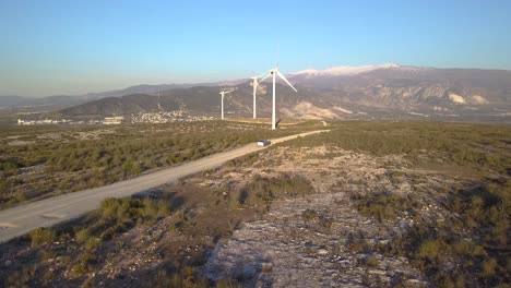 Aerial-view-of-a-car-driving-off-road-in-the-countryside-surrounded-by-wind-mills-and-mountains-on-the-background