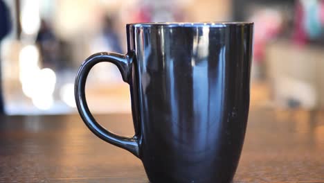 In-Einem-Café-Aufgenommener-Clip-Einer-Schwarzen-Und-Braunen-Kaffeetasse-Mit-Heißem-Kaffee-Oder-Tee-Darin,-Während-Sich-Im-Hintergrund-Menschen-Bewegen
