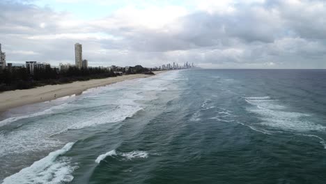 Slow-aerial-footage-of-a-beach-at-sunrise-with-white-sand,-wave-rolling,-swimmers-surfers-and-nice-beachfront-with-numerous-towers-and-buildings