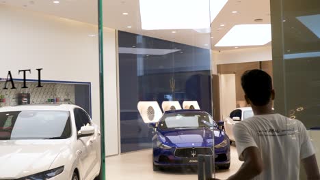 Following-young-Asian-male-through-glass-showroom-doors-to-view-Maserati-dealership-in-Luxury-shopping-mall,-Icon-Siam,-Bangkok,-Thailand