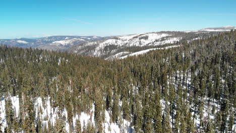 Aerial-shot-of-snow-covered-mountains-and-forest,-camera-flies-forward-and-tilts-up-slowly-revealing-panoramic-views-of-the-Sierra-Nevadas
