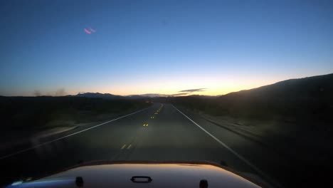 Conduciendo-Hacia-El-Amanecer-En-Un-Vehículo-Rojo-En-El-Desierto-Con-La-Luz-De-Los-Faros-Y-Las-Montañas-Visibles-En-Silueta