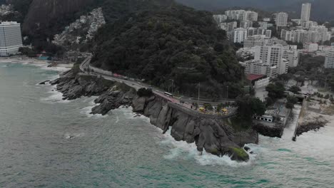 Aerial-approach-on-Leblon-viewpoint-and-parking-with-waves-crashing-in-on-the-rocky-coast-on-a-gray-overcast-day-in-Rio-de-Janeiro