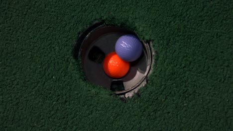 Eine-Nahaufnahme-Eines-Orangefarbenen-Balls-In-Einem-Loch-Aus-Der-Vogelperspektive,-Und-Dann-Fallen-Ein-Violetter-Und-Ein-Gelber-Golfball-übereinander-In-Das-Golfloch