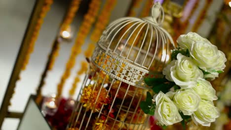 White-roses-and-marry-gold-flowers-bouquet-set-in-a-cage-with-Candles-in-glasses-and-rose-petals-on-the-table,-Rotational-Panning-view