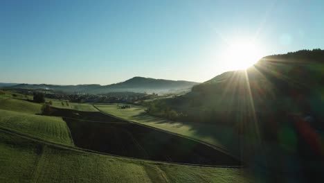 Luftdrohnenansicht-Der-Morgenstunde-über-Friedlicher-Landschaft-Mit-Grünen-Und-Landwirtschaftlichen-Feldern,-Sonne-Scheint-Direkt-In-Die-Kamera,-Schweiz
